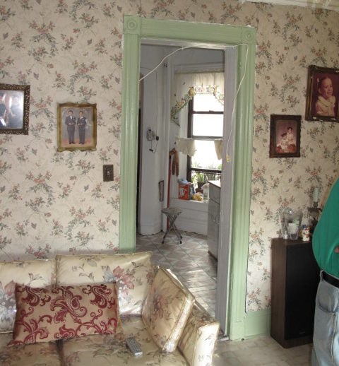 Color photo of the Saez-Velez family's living room with pictures hanging up on floral wallpaper that matches the plastic-covered couch