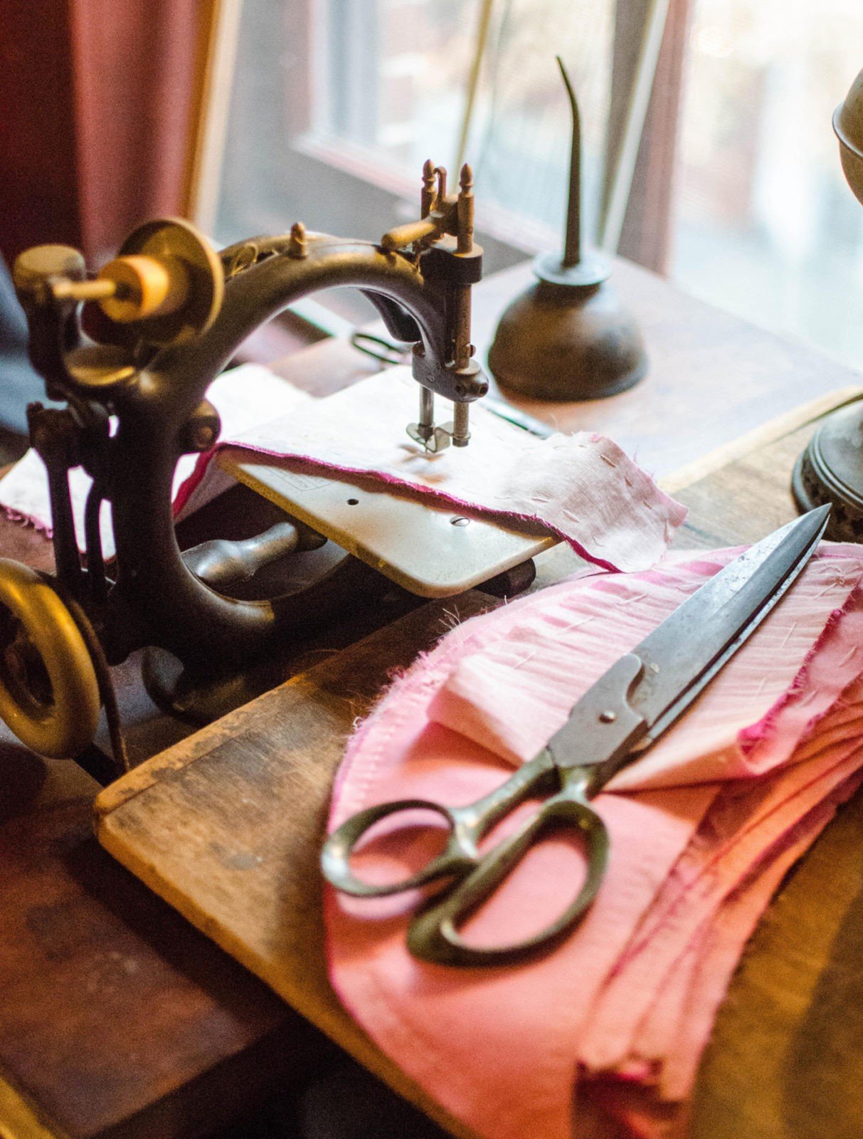 A strip of pink fabric is between the needle and base of an old wooden sewing machine next to a pair of metal scissors