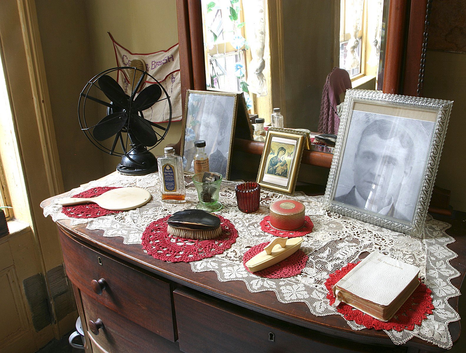 Brown vanity dresser with doilies, a fan, products, hair brushes, and photos of two family members inside the recreated Baldizzi apartment