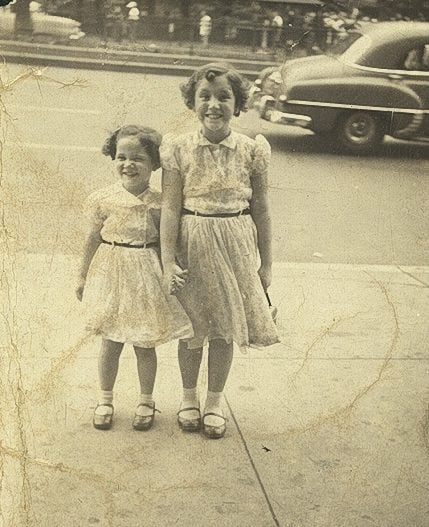 Bluma and Bella in matching dresses, holding hands and smiling on the sidewalk