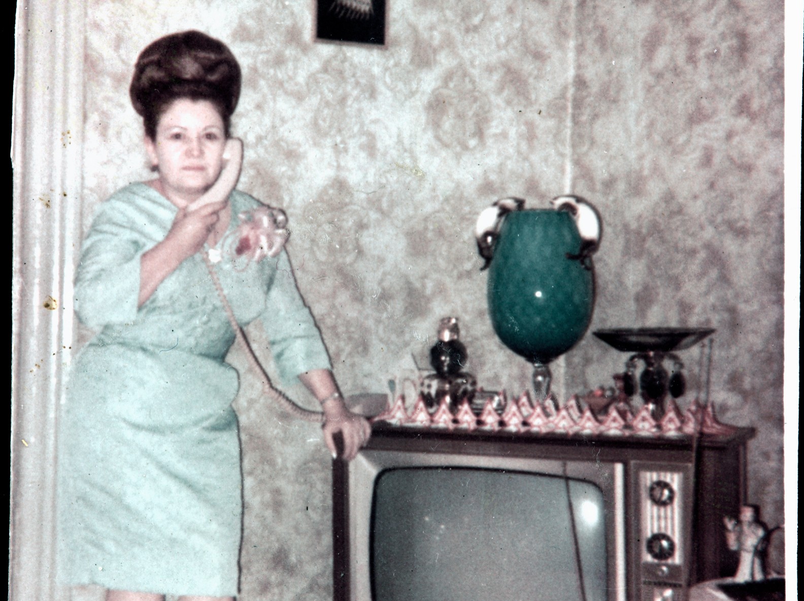 Ramonita Saez in a pastel green dress with a beehive hairstyle stands next to a TV while speaking on the phone.