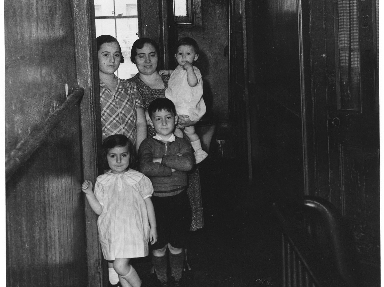 Adult carrying a baby while standing beside an older child and 2 younger children in a doorway in the hallway of a tenement