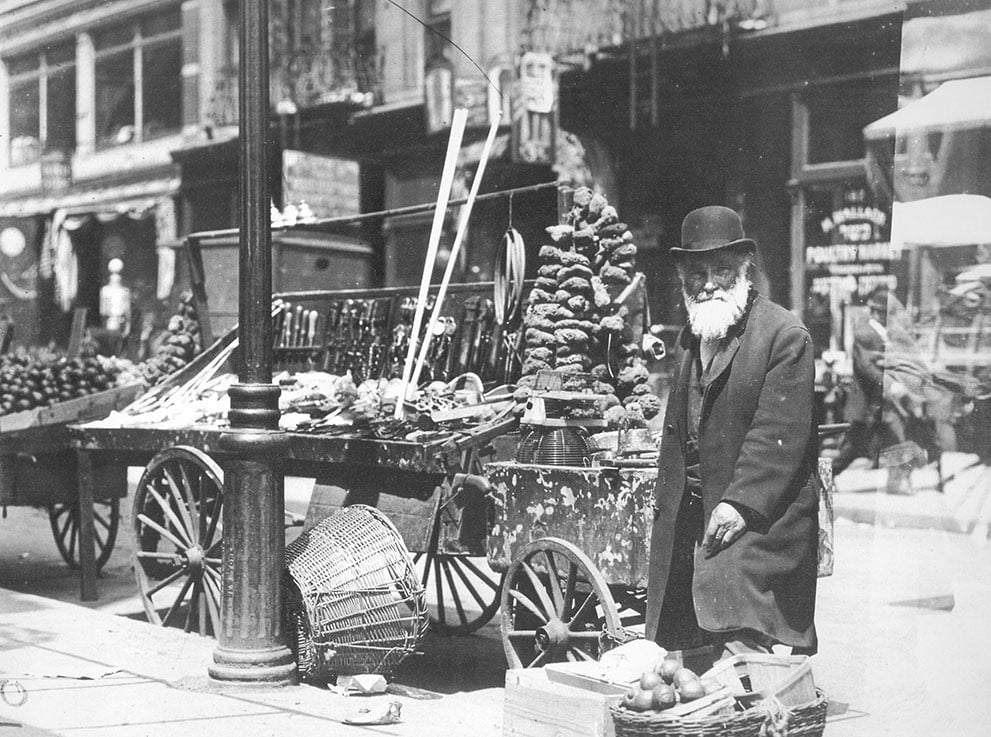 Black and white photo of a bearded street vendor in a hat and coat standing beside their pushcart full of goods