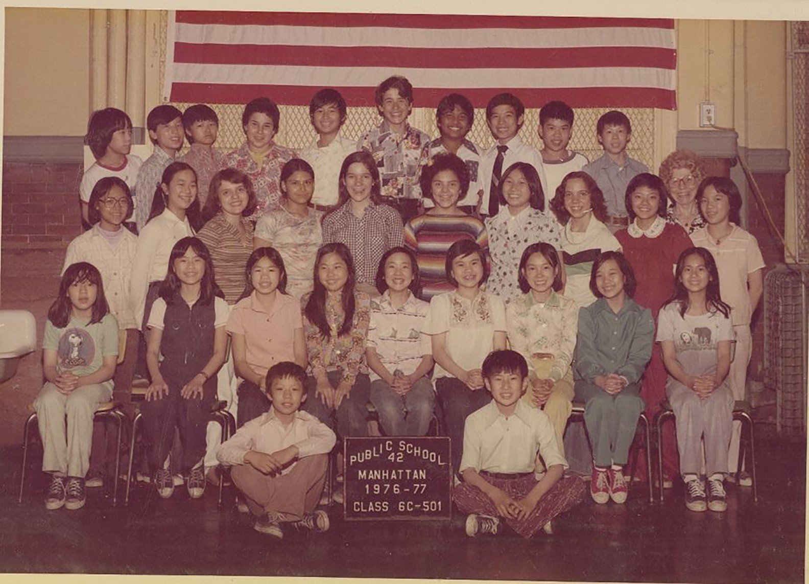 A diverse class of 31 students and one teacher. A sign in front reads, “Public School 42, Manhattan 1976-77, Class 6c-501.”