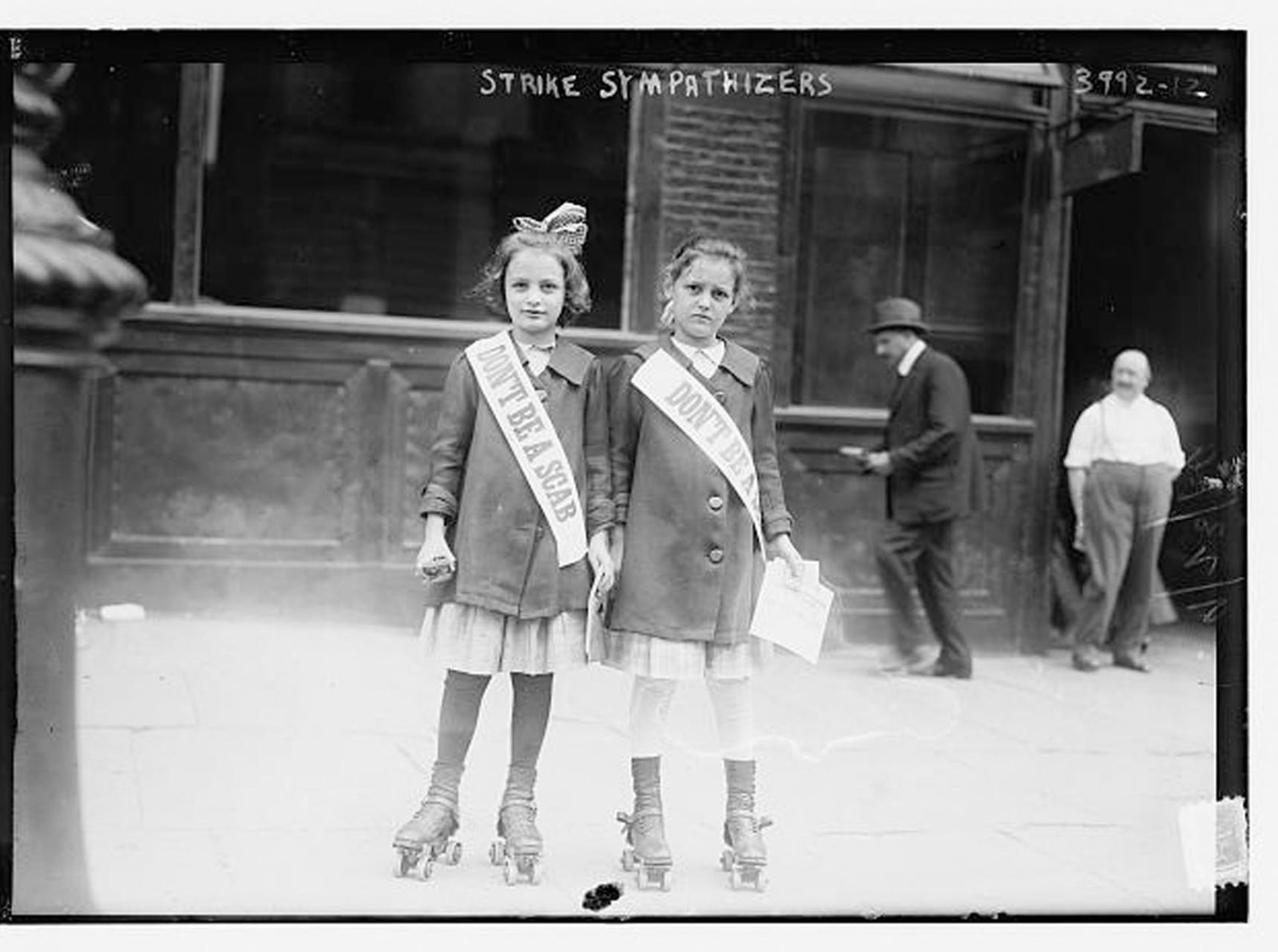 Two young girls side by side in roller skates and matching jackets and skirts wear strike sashes that say "Don't be a scab"