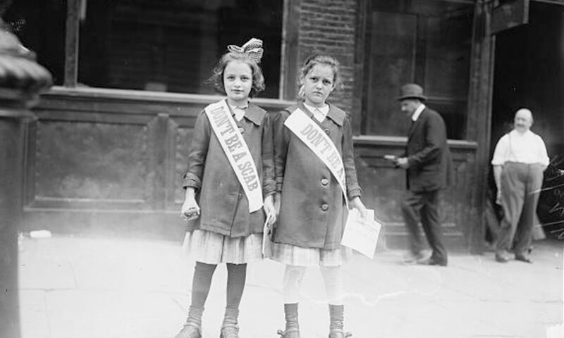 Two young girls side by side in roller skates and matching jackets and skirts wear sashes that say "Don't be a scab"