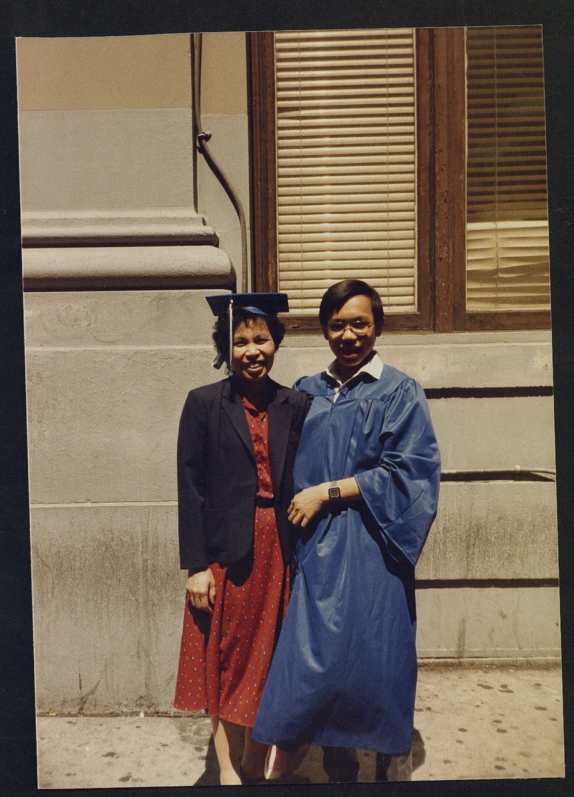 Kevin, in a blue graduation gown, stands with his mother who is wearing his graduation cap and a red dress and navy blazer.