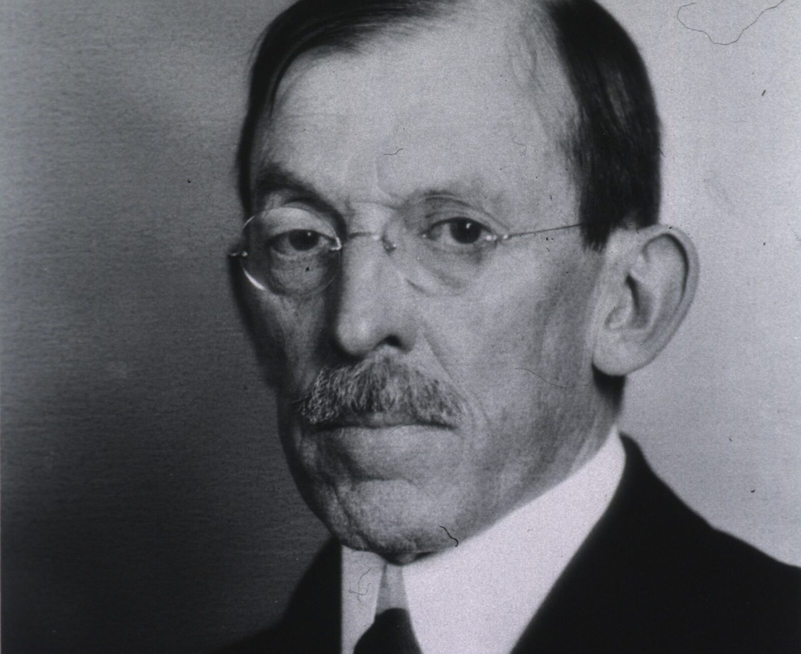 Black and white photo of an older man with a receding hairline, mustache, rimless glasses, and a serious expression marked by a raised eyebrow