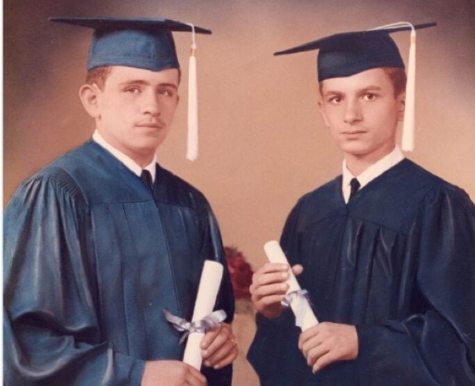 Two young men with light skin tones stand opposite each other dressed in dark blue graduation caps and gowns and holding rolled-up diplomas