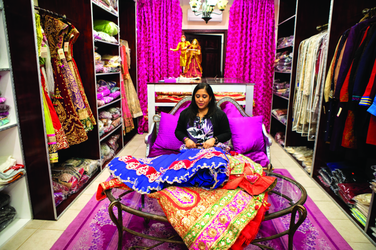 Julie sits on a couch in her shop. In front of her, on racks to either side, are colorful garments with embroidery. 