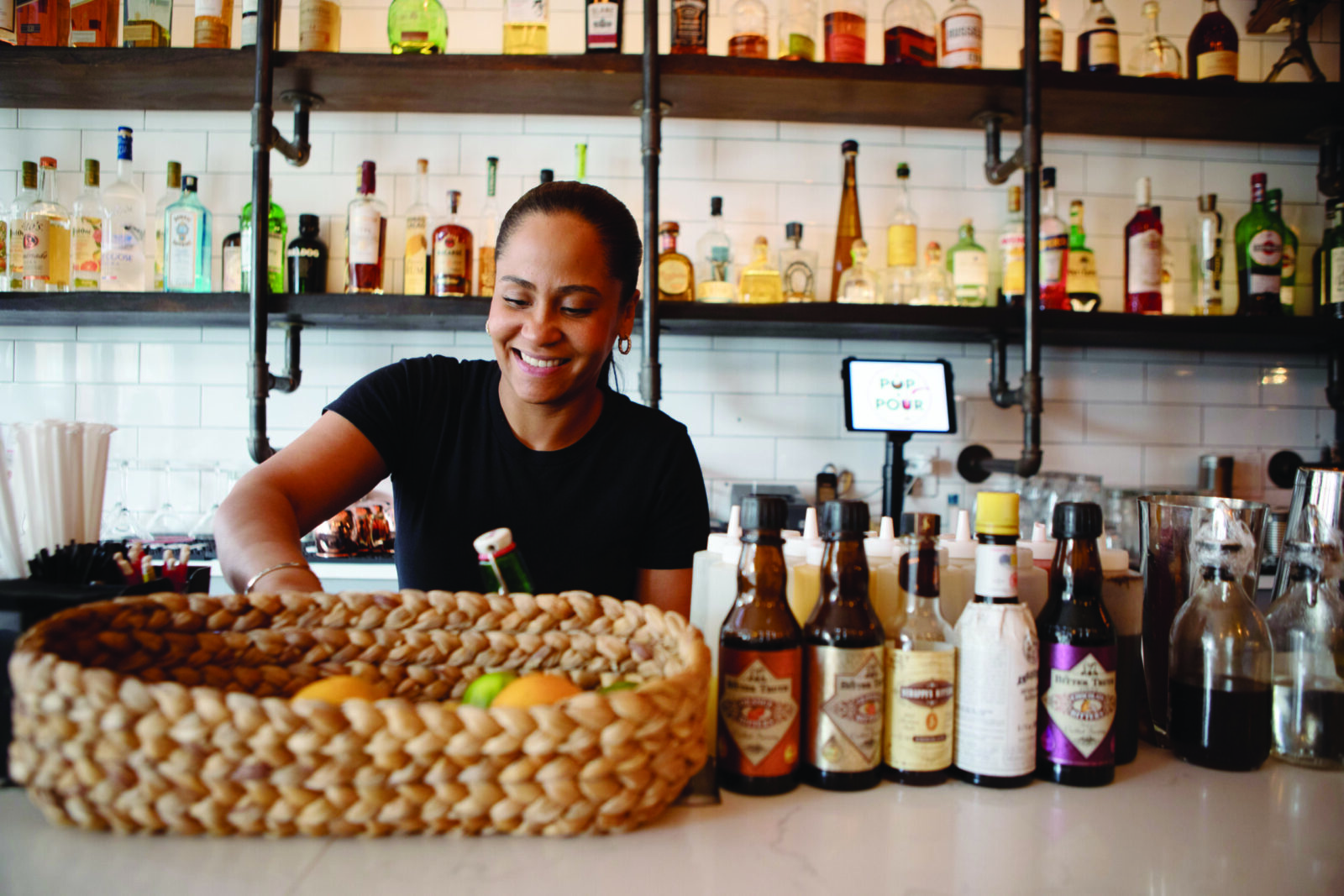 Yajaira smiles down as she works behind the bar, her hands obscured by a basket of fruit. Liquor bottles line the shelves behind her.