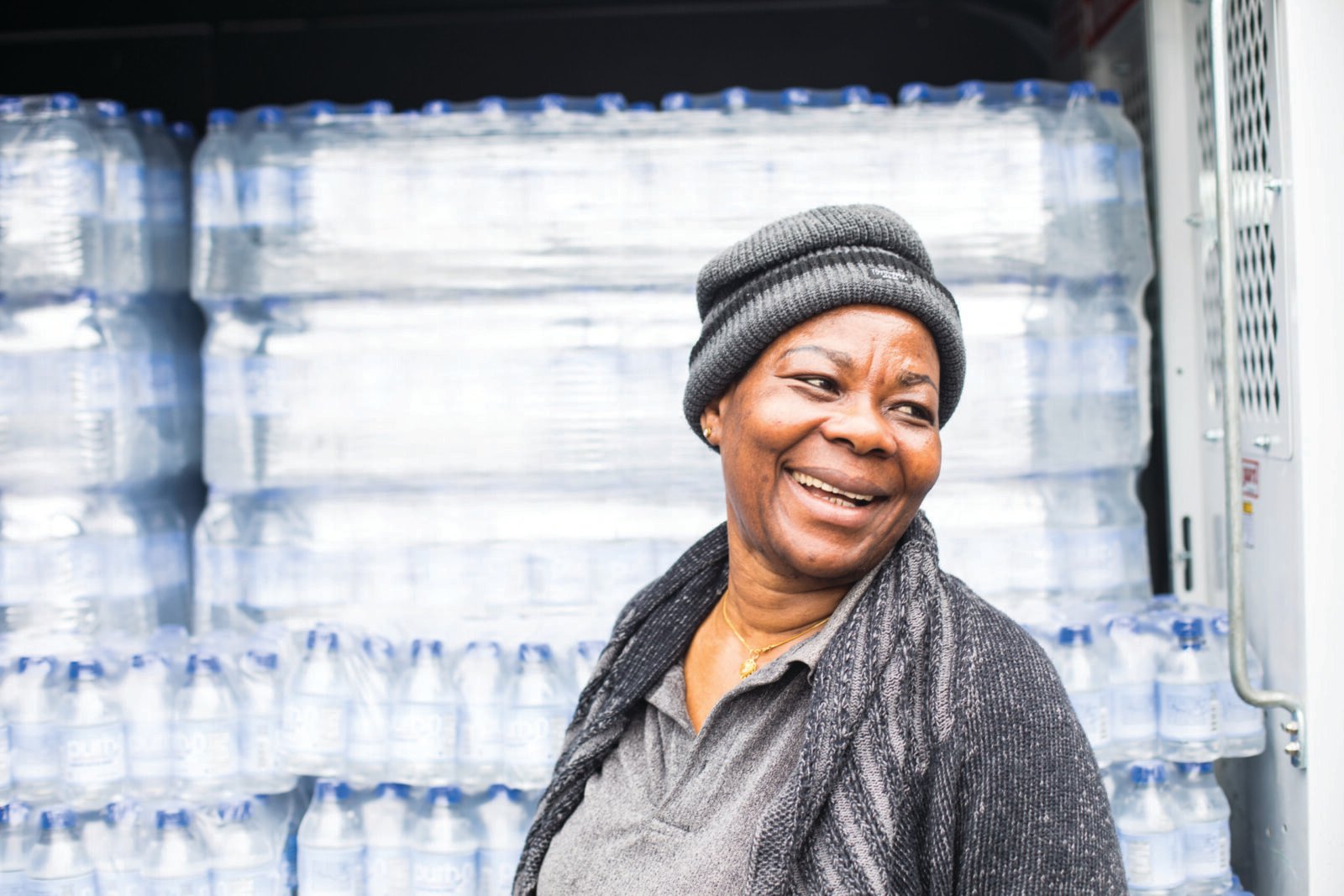 Atinuke, a woman with dark skin in a gray hat and clothes, smiles off to the right. She is standing in front of a wall of plastic water bottles in cases. 