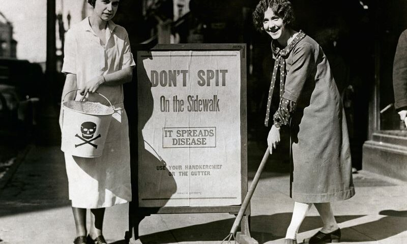 Two women mop up a spot on the sidewalk where someone expectorated by an anti-spitting sign during a public health campaign in Syracuse, New York, in 1900