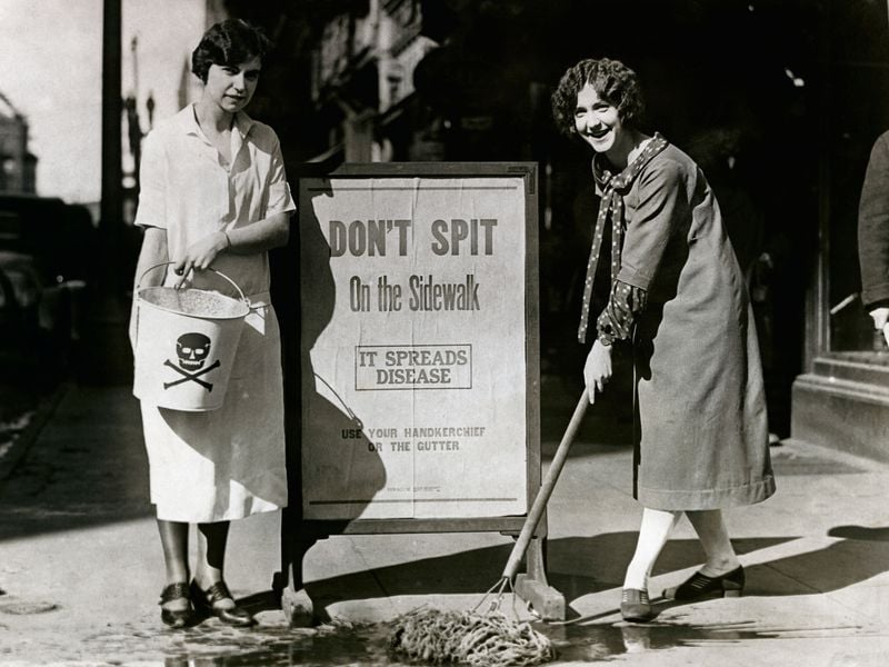 Two women mop up a spot on the sidewalk where someone expectorated by an anti-spitting sign during a public health campaign in Syracuse, New York, in 1900