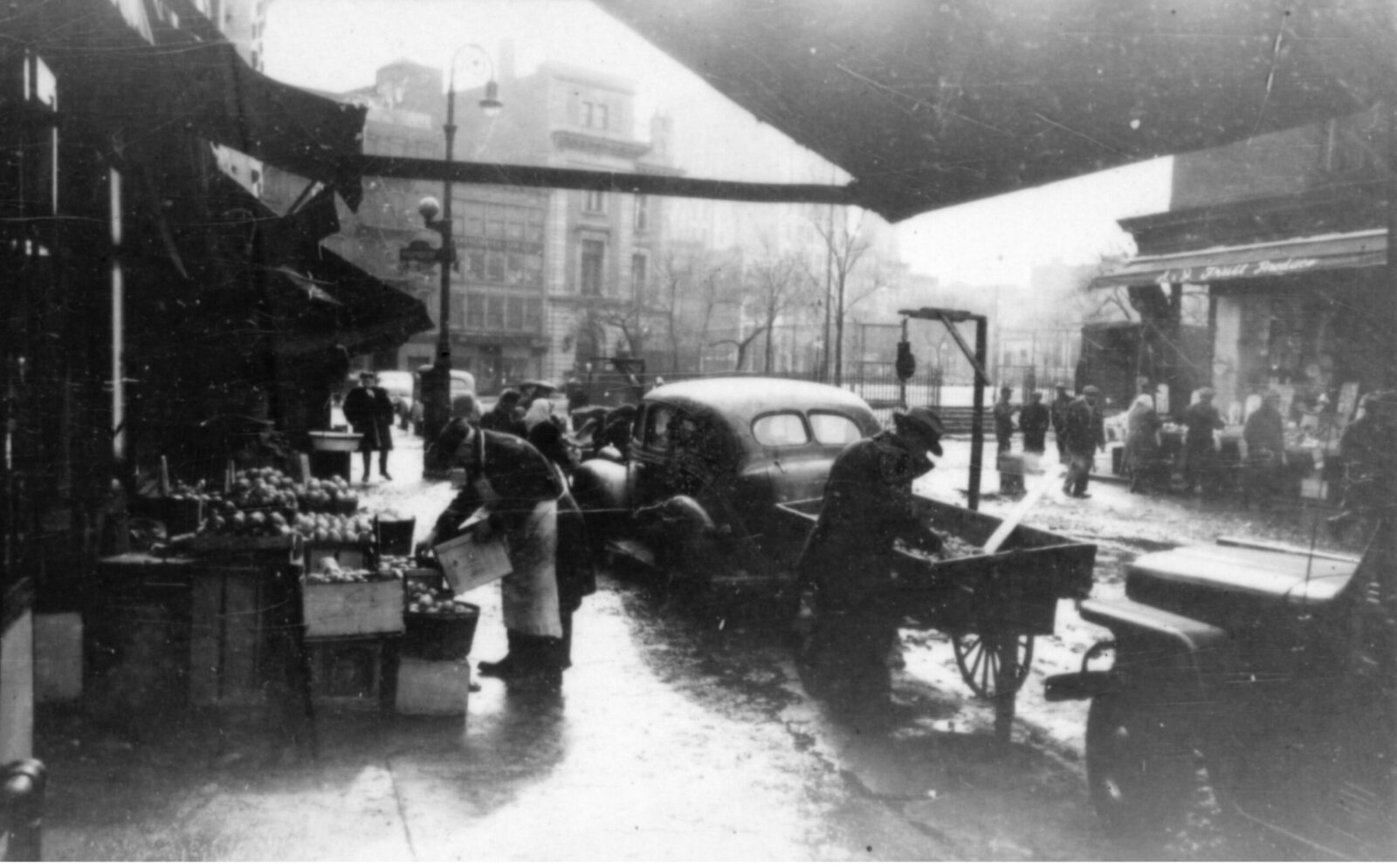 Black and white photo of the sidewalk in front of a food stand. A man in an apron is stooped over a fruit display. Another man in a hat and coat is bent over a push cart between two old cars.