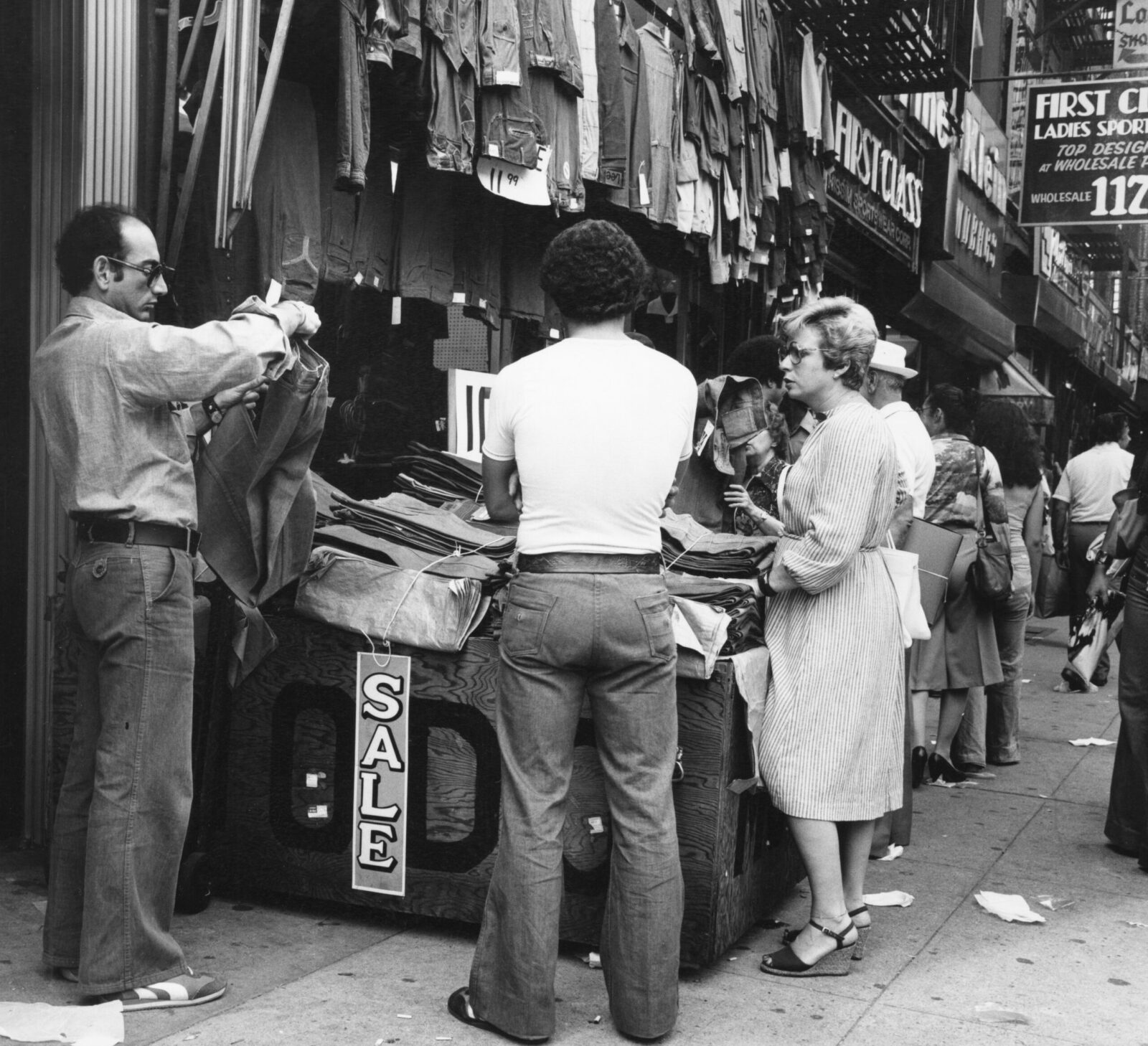 Black and white photograph of people shopping for pants outside the storefront of a Lower East Side clothing store. Tables sit on the sidewalk with merchandise.