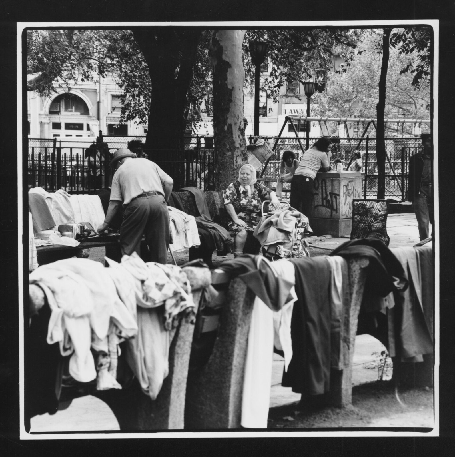 Black and white photo of clothing being sold on benches in Seward Park. An older woman watches a man browse clothing while children swing on a playground in the background.