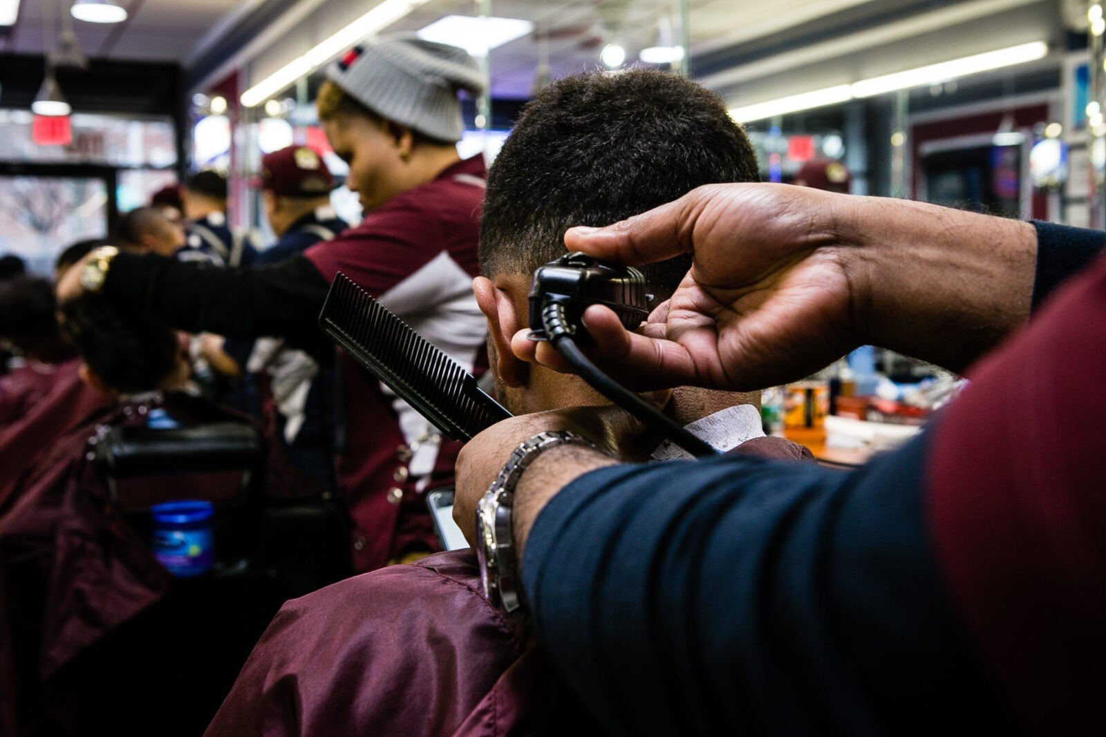 Close up of a customer  getting their hair cut. The barber is using clippers and a plastic comb. Their reflection can be seen in the mirror.  