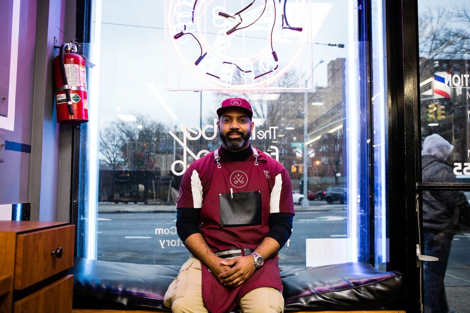 Nestor seated at the front window of his shop wearing a maroon hat and apron with his logo, open scissors and with the letters “T,” “B,” and “F”. 