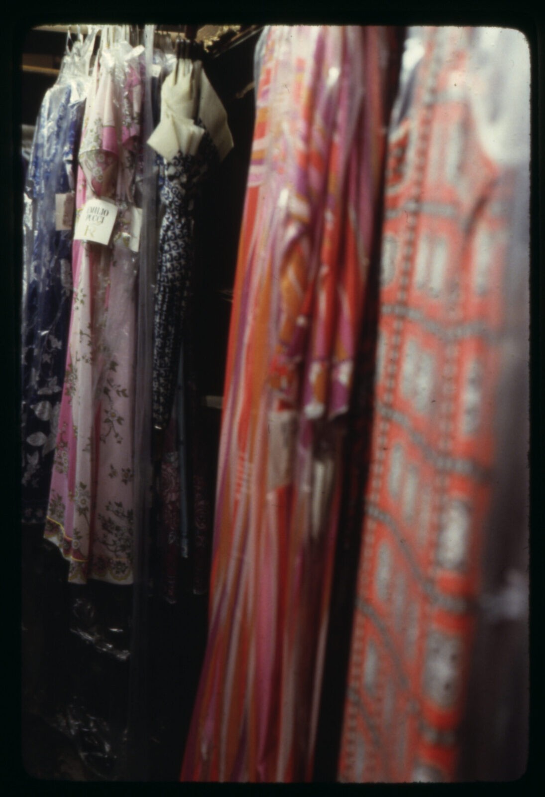 Four colorful garments with 1970s patterns wrapped in plastic hanging from racks. A blue and white umbrella also hangs from one of the racks.