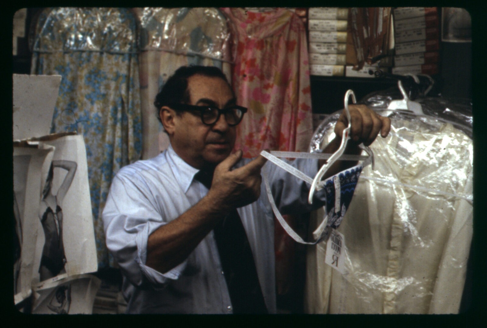 Sidney, a man with light colored skin and dark hair wears black glasses, a shirt, and tie. Surrounded by colorful garments he holds thong underwear.
