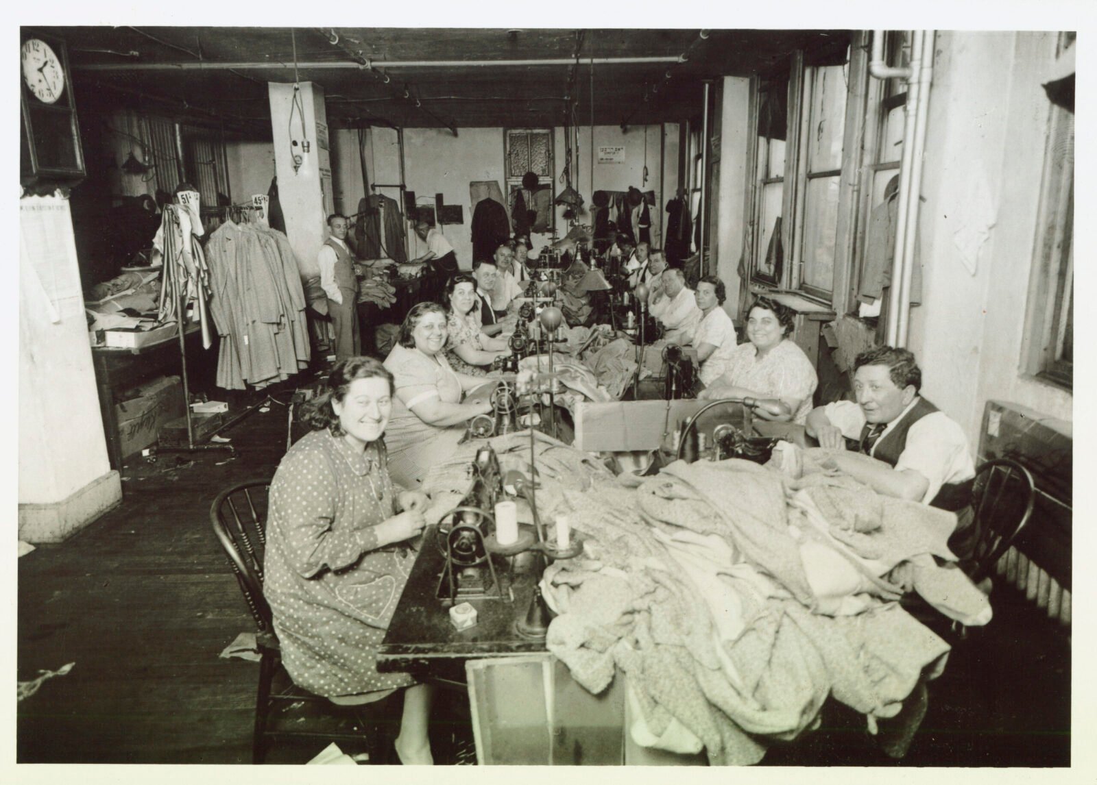 Black and white photograph, 13 people with light skin seated at a long table with sewing machines, garments. Rosaria has dark hair and is smiling.