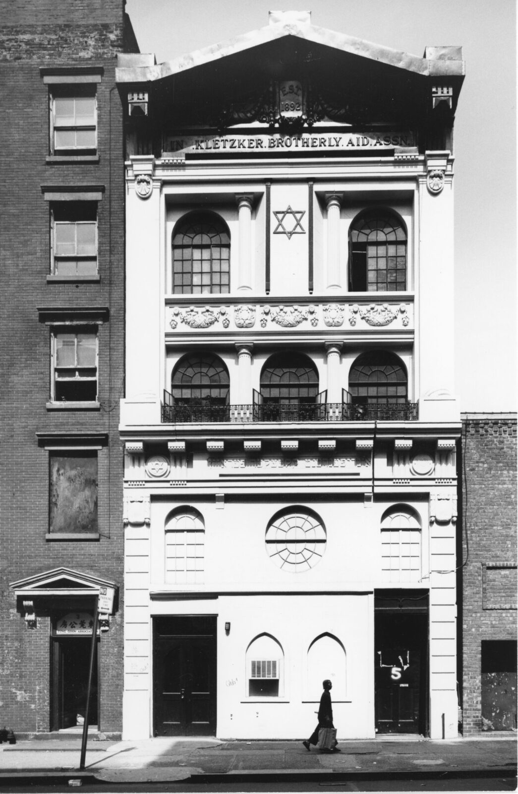 Black and white photograph of a four story tall building. It is adorned with a Jewish star and a plaque near the top “Kletzker Brotherly Aid Assn.” 