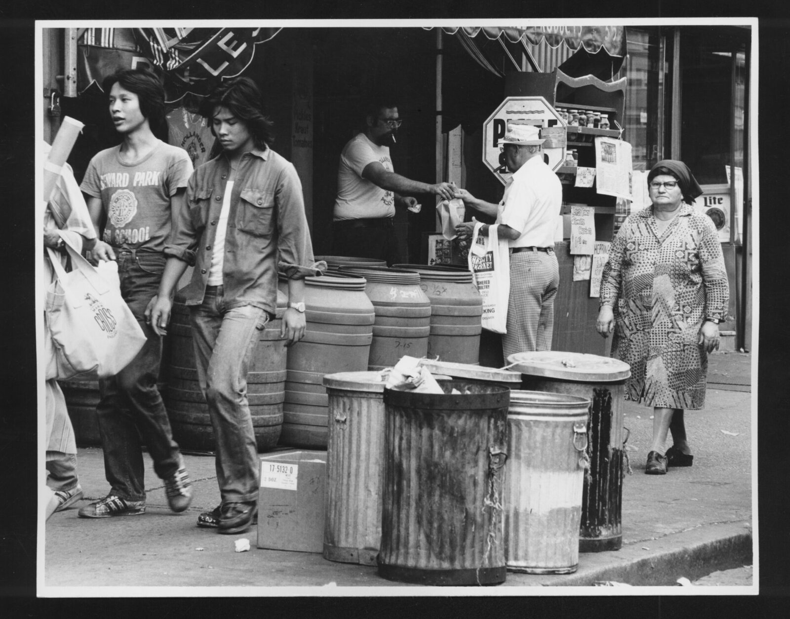 Black and white photo of the sidewalk in front of L. Hollander and Sons pickle shop. Two young adults walk past an older adult picking up a purchase on a street lined with trash bins and pickle barrels