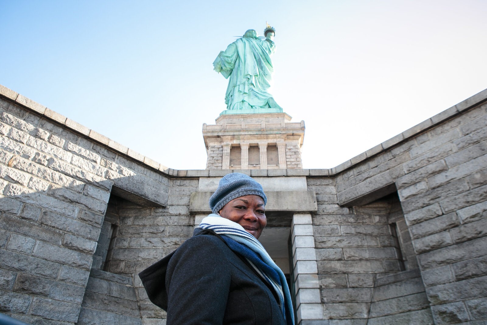 Atinuke stands smiling at the camera at the base of the Statue of Liberty. She is dressed for winter, the Statue of Liberty facing backwards.