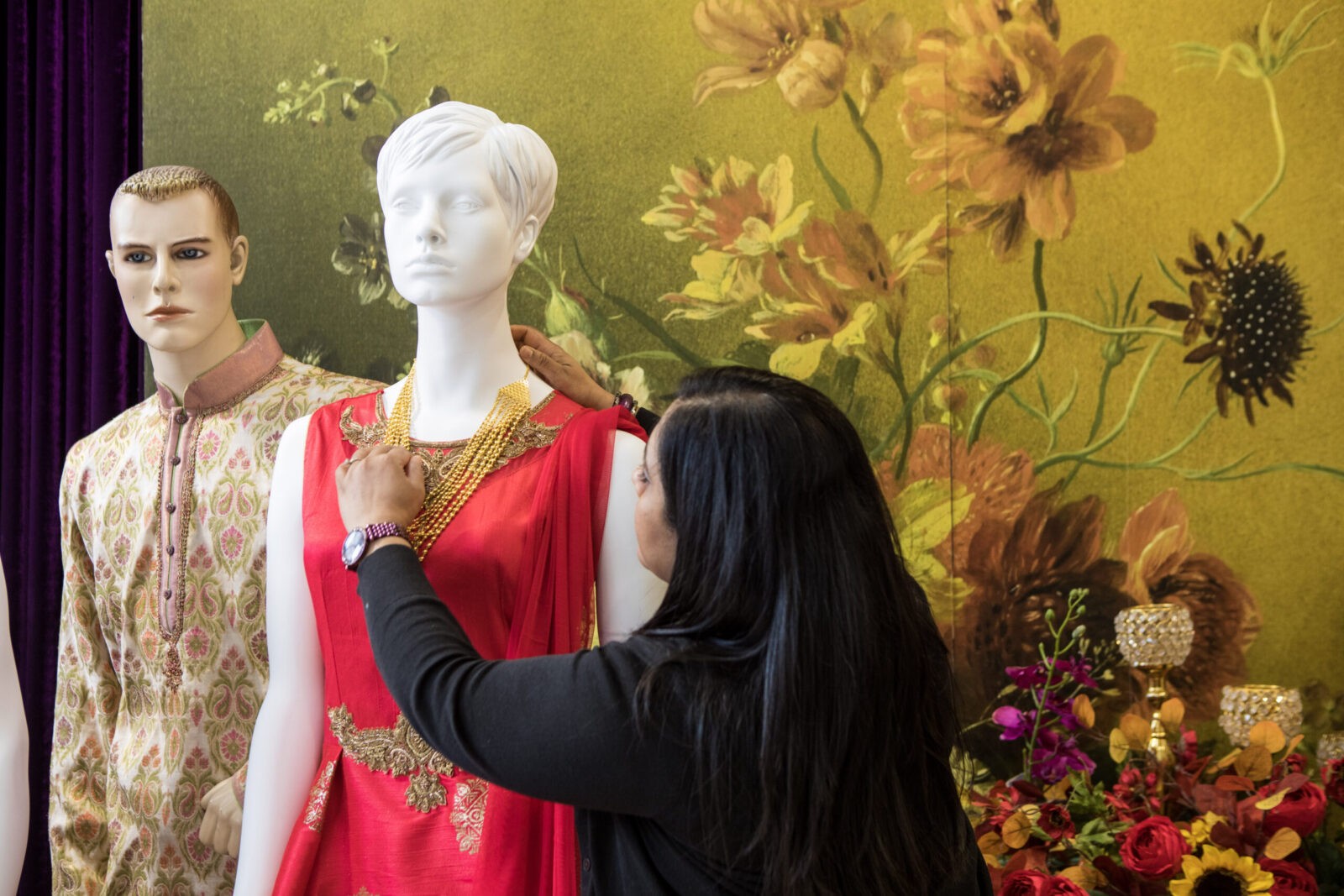 Julie, with medium skin and dark hair, styling a mannequin in a a red sleeveless dress with gold embroidery with a gold necklace. Another mannequin wears a floral patterned kurta.
