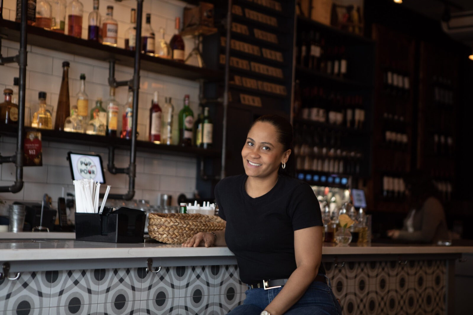 Yajaira sits in front of the bar and smiles at the camera. On the bar is a basket and a container of straws. 